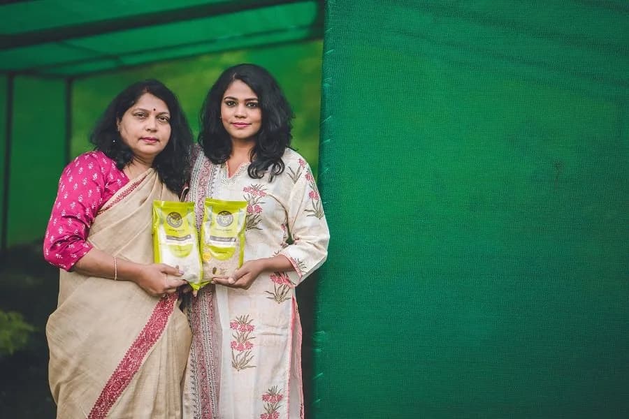Two women in saris holding packets of a food product against a green backdrop
