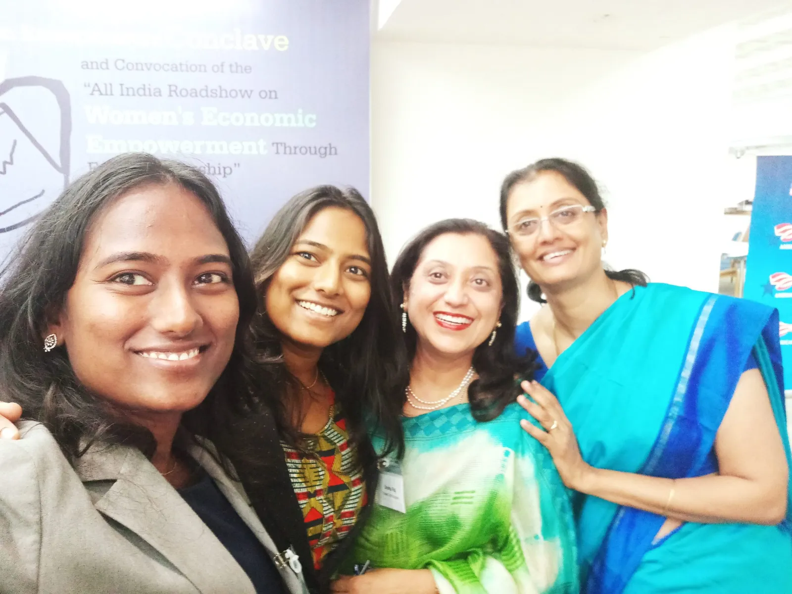 Four women in formal dress smiling together in front of an event banner