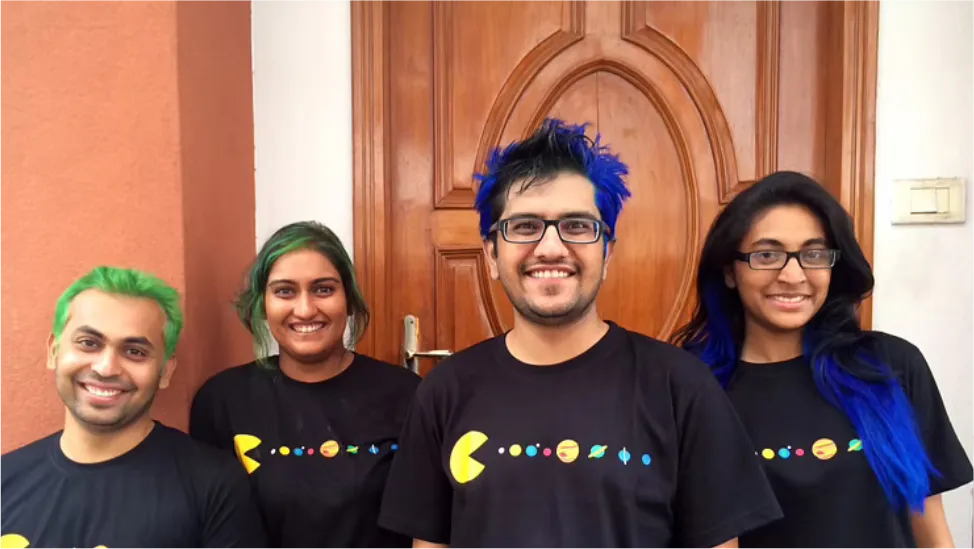 Four people with brightly dyed hair in matching printed T-shirts by a wooden door