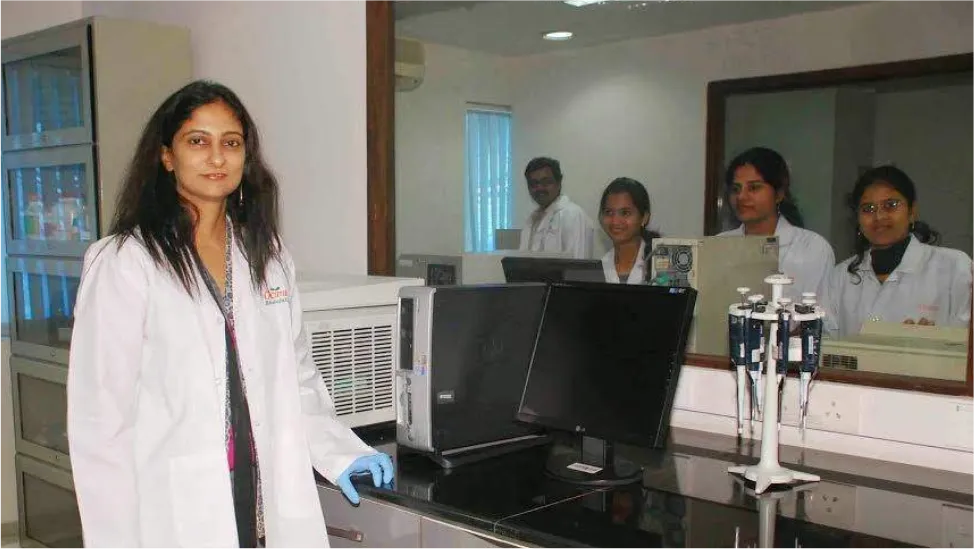 Person in a white coat beside laboratory instruments, others standing behind glass